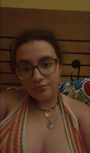 Woman with glasses in halter top and necklaces in bedroom.