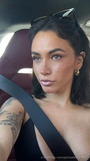 Woman with dark hair and tattoos in a black top, sitting in a car with seatbelt on.