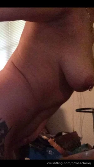 Woman with tattoo on arm, bare chest, nipple piercing, indoors.
