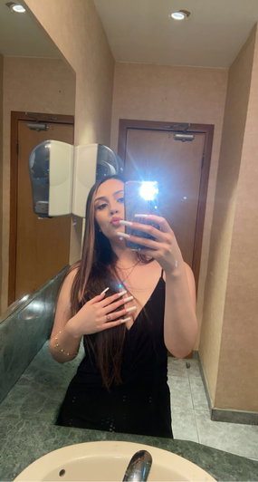 Woman with long hair in black dress taking bathroom mirror selfie.