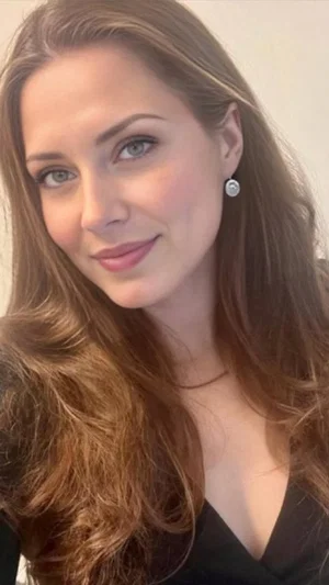 Woman with long brown hair in black top, indoor selfie, smiling softly.