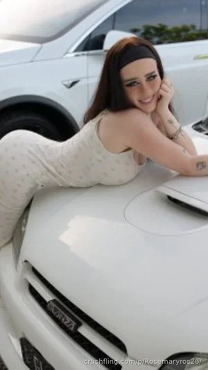 Woman in light patterned dress lying on white car hood, smiling, outdoor setting.