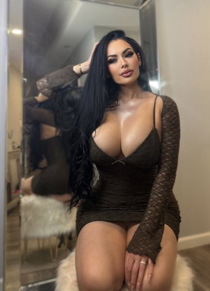 Woman with long black hair in dark lace dress posing in front of mirror indoors.