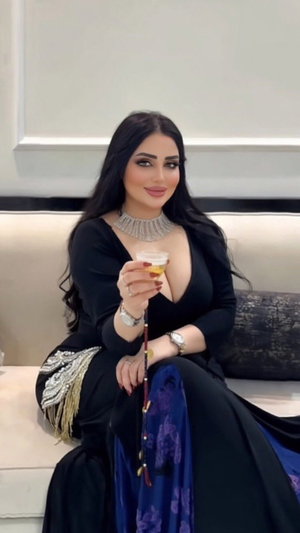 Woman in black dress with plunging neckline holding drink, sitting on couch indoors.