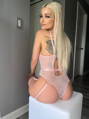 Woman with blonde hair in pink sheer lingerie posing on white stool indoors.