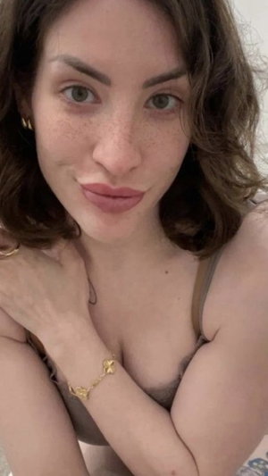 Woman with wavy brown hair and freckles wearing a light-colored bra, posing indoors.