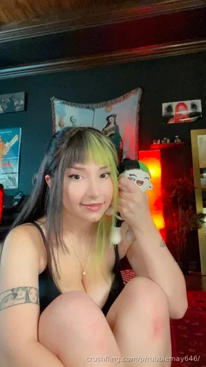 Woman with green-tipped hair in black top, sitting indoors with plush toy, tattoos visible.