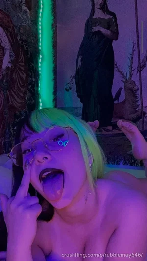 Woman with green hair and glasses, topless, posing in front of a mystical-themed backdrop with neon
