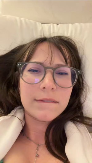 Woman with glasses lying on bed, wearing necklace, partially covered by white sheets.