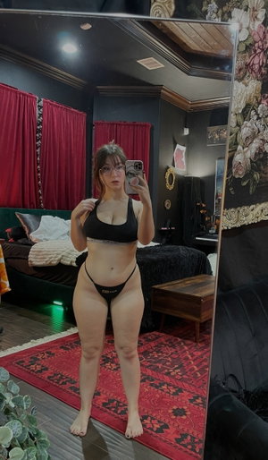 Woman in black lingerie taking mirror selfie in bedroom with red curtains and rug.