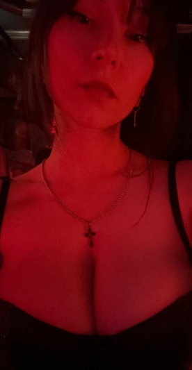 Woman with dark hair in black top under red lighting, wearing cross necklace.
