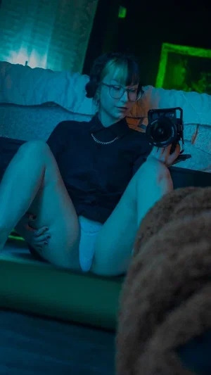 Woman with glasses in black top and white underwear sits on couch holding camera, dimly lit room