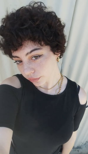 Woman with curly dark hair in black cold-shoulder top, wearing gold necklace, indoors.