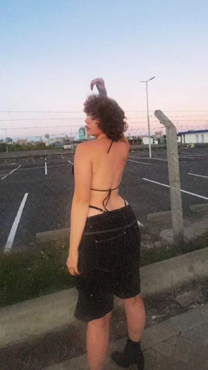 Woman in black strappy top and skirt standing in parking lot at dusk.