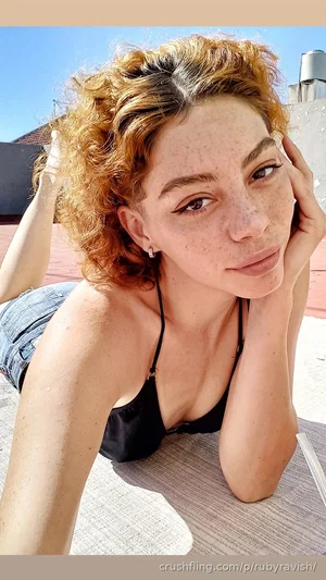 Woman with curly hair in black top and denim shorts lying on outdoor surface.