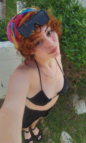 Woman with curly red hair in black bikini top and shorts, wearing colorful headwrap, outdoors on