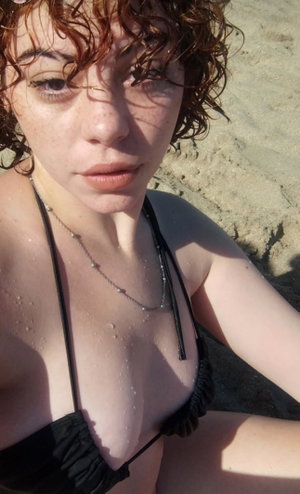 Woman with curly hair in black bikini top on sandy beach, wearing necklace.