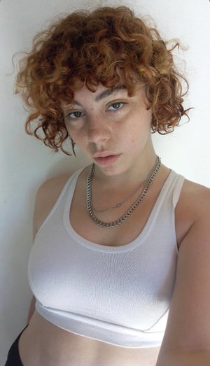 Woman with curly red hair wearing white crop top and chain necklace, standing indoors.