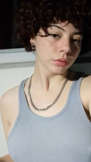 Woman with curly hair wearing a gray tank top indoors, looking at camera.
