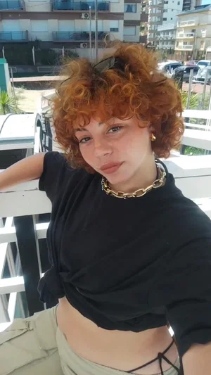 Woman with curly red hair in black crop top and beige pants, sitting outdoors on balcony.