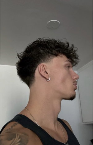 Man with dark curly hair, earring and tattoo in black tank top indoors.