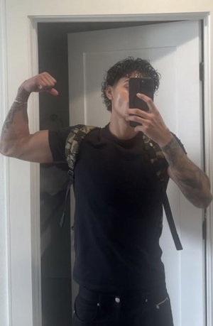 Tattooed man with curly hair flexing in black t-shirt indoors.