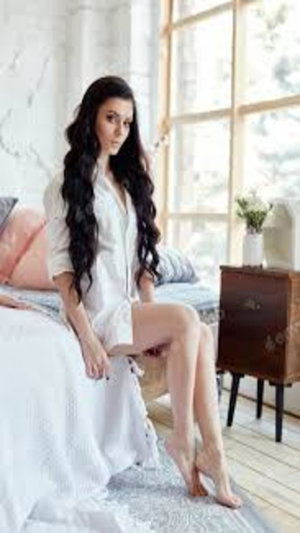 Woman with long dark hair in white dress sits on bed.