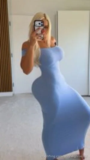 Blonde woman in a blue off-shoulder dress taking a mirror selfie indoors.