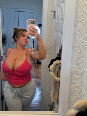 Woman in pink top and gray sweatpants takes mirror selfie in bedroom.