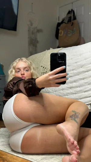 Blonde woman in white underwear and brown top takes mirror selfie on bed.