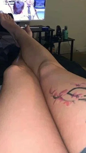 Woman with tattoo on leg, legs crossed, watching TV in bedroom.