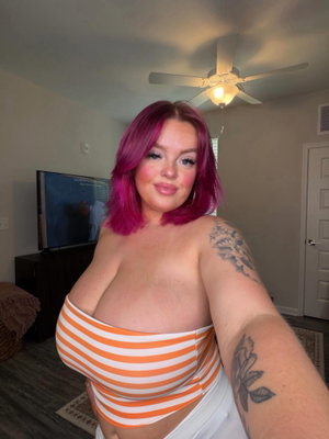 Woman with pink hair and tattoos wears orange-white striped top in bedroom.
