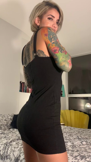Woman with short blonde hair and colorful arm tattoos wears black dress in bedroom.