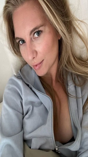 Blonde woman in gray zip-up hoodie taking a selfie indoors.