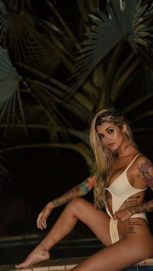 Tattooed blonde woman in white swimsuit posing outdoors at night.