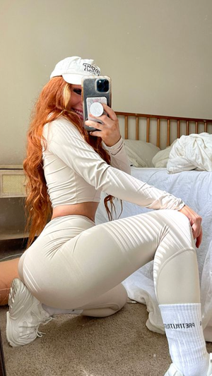 Woman with long red hair in white crop top and leggings, kneeling in bedroom taking mirror selfie.