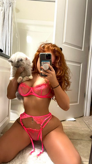 Woman with red curly hair in pink lingerie holding a cat, kneeling in a bathroom.