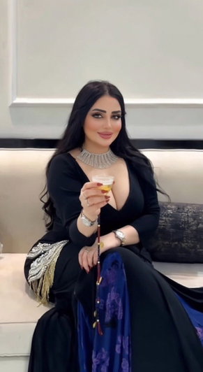 Woman in black dress with deep neckline, holding drink, sitting on couch indoors.