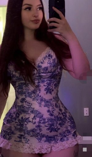 Woman with long dark hair in floral dress taking mirror selfie indoors.