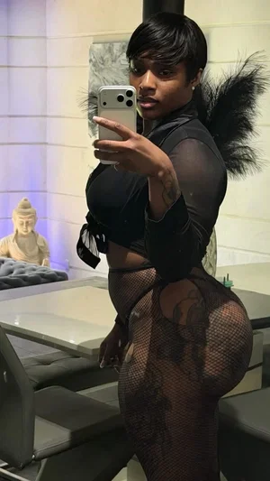 Woman in black sheer outfit with feathered backpiece, taking mirror selfie in modern room.