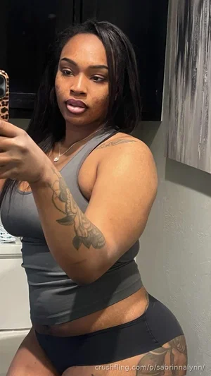 Woman with tattoos in gray top and black bottoms, taking mirror selfie indoors.