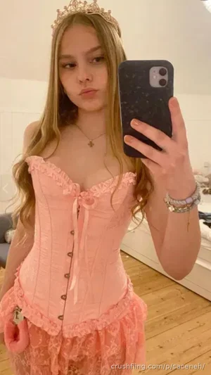 Woman in pink corset and tiara taking mirror selfie in bedroom.