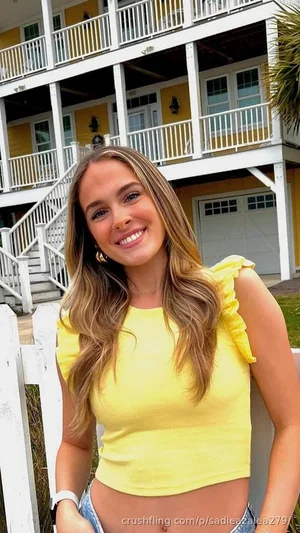 Woman with long brown hair in yellow crop top and jeans, smiling outdoors in front of yellow house.