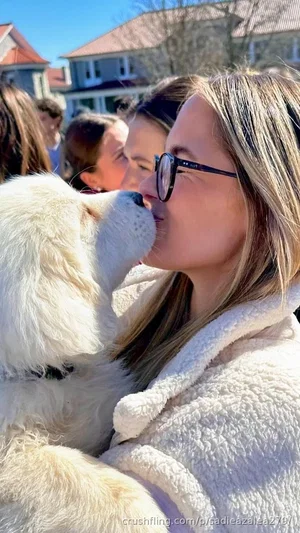 Woman with blonde hair and glasses kissing a fluffy white dog outdoors.