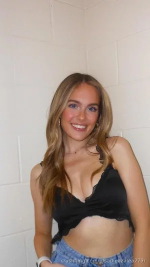 Woman with blonde hair in black crop top and jeans, smiling in front of white tiled wall.