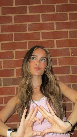 Woman with long wavy hair in a pink top making a heart shape with her hands against a brick wall.