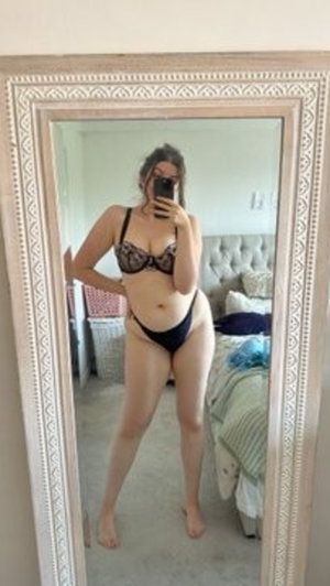 Woman in black lace bra and underwear taking mirror selfie in bedroom.