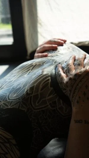 Person with full back and arm tattoos lying near window, hands resting on back.