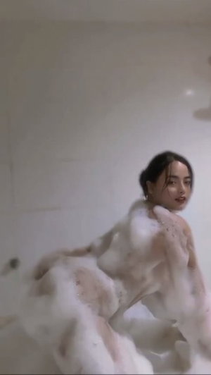 Woman covered in foam in a bathroom, facing camera with dark hair pulled back.