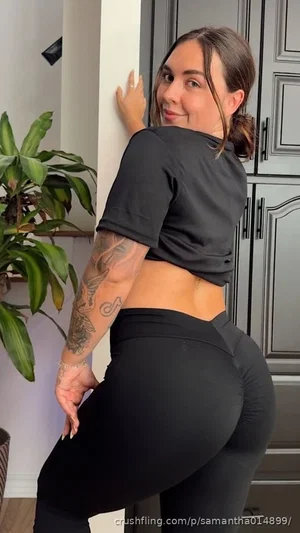 Woman in black crop top and leggings, posing indoors with tattooed arm, near plant and cabinets.